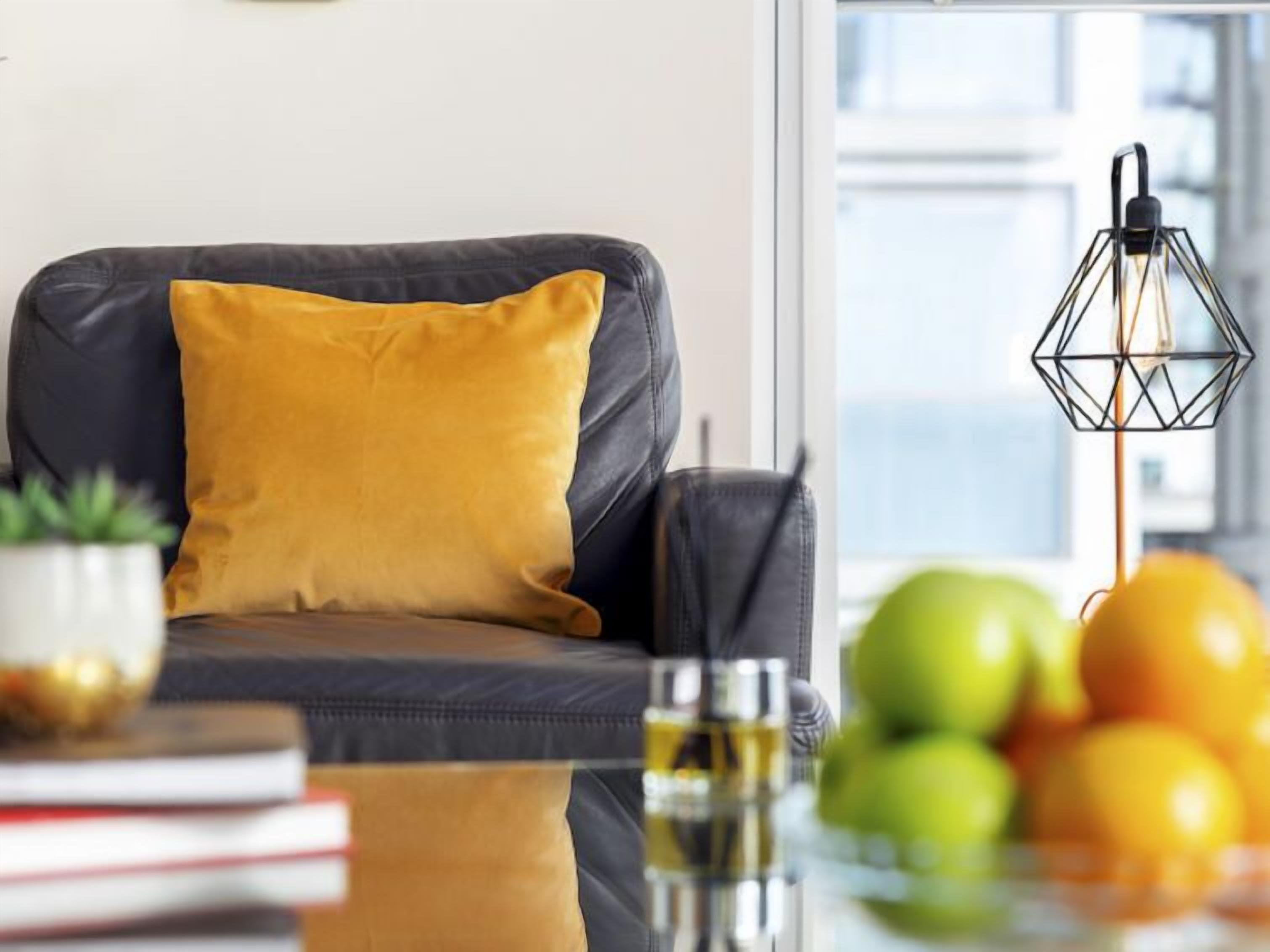 Ein modernes Wohnzimmer mit einem dunklen Sofa, einem gelben Kissen, einem Glastisch mit einem Getränk und einer Obstschale.
