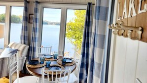 Uma área de refeições com uma mesa posta para quatro pessoas, grandes janelas com cortinas azuis e uma placa com a inscrição 'LAKE'.
