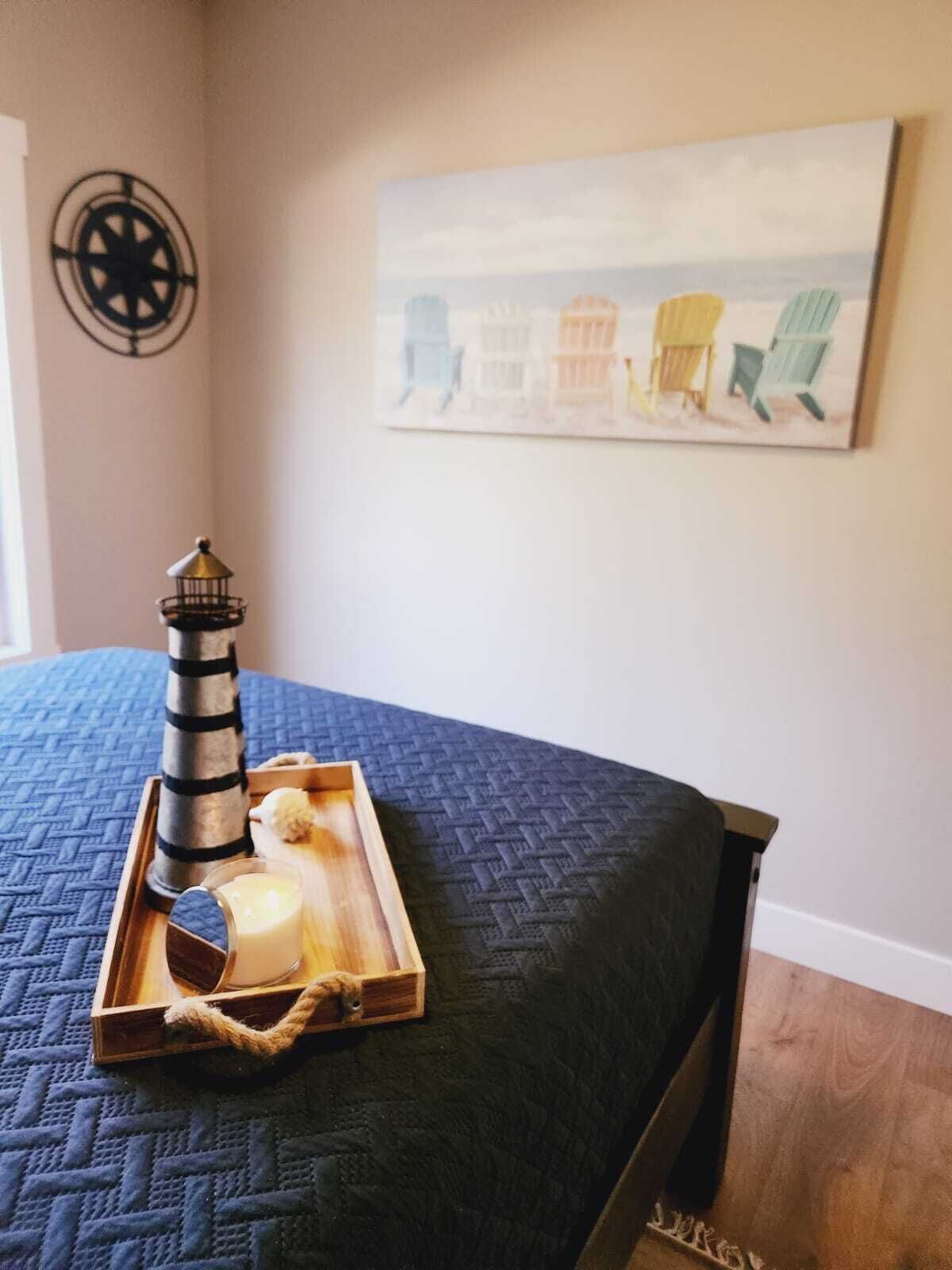A neatly made bed with a blue quilt, a wooden tray with a lighthouse model, and a painting of beach chairs on the wall.