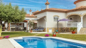 Un espace aménagé au bord d’une piscine à l’eau d’un bleu limpide, un coin salon extérieur abrité et un bâtiment doté d’un toit en tuiles et de détails en pierre.