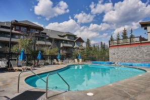 A swimming pool with lounge chairs and umbrellas, surrounded by buildings with stone walls and a clear sky.