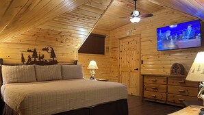Tunnelmallinen hirsihuone, jossa on sänky, lipasto ja televisio, josta näkyy jokin ohjelma.