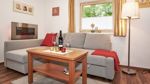 Un salon avec un canapé gris, une table basse en bois, un téléviseur fixé au mur, une fenêtre avec des rideaux et un lampadaire.