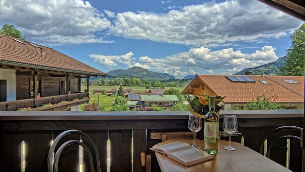Une terrasse offrant une vue sur un paysage montagneux, avec des meubles en bois et une table sur laquelle se trouvent une bouteille de vin et un verre.