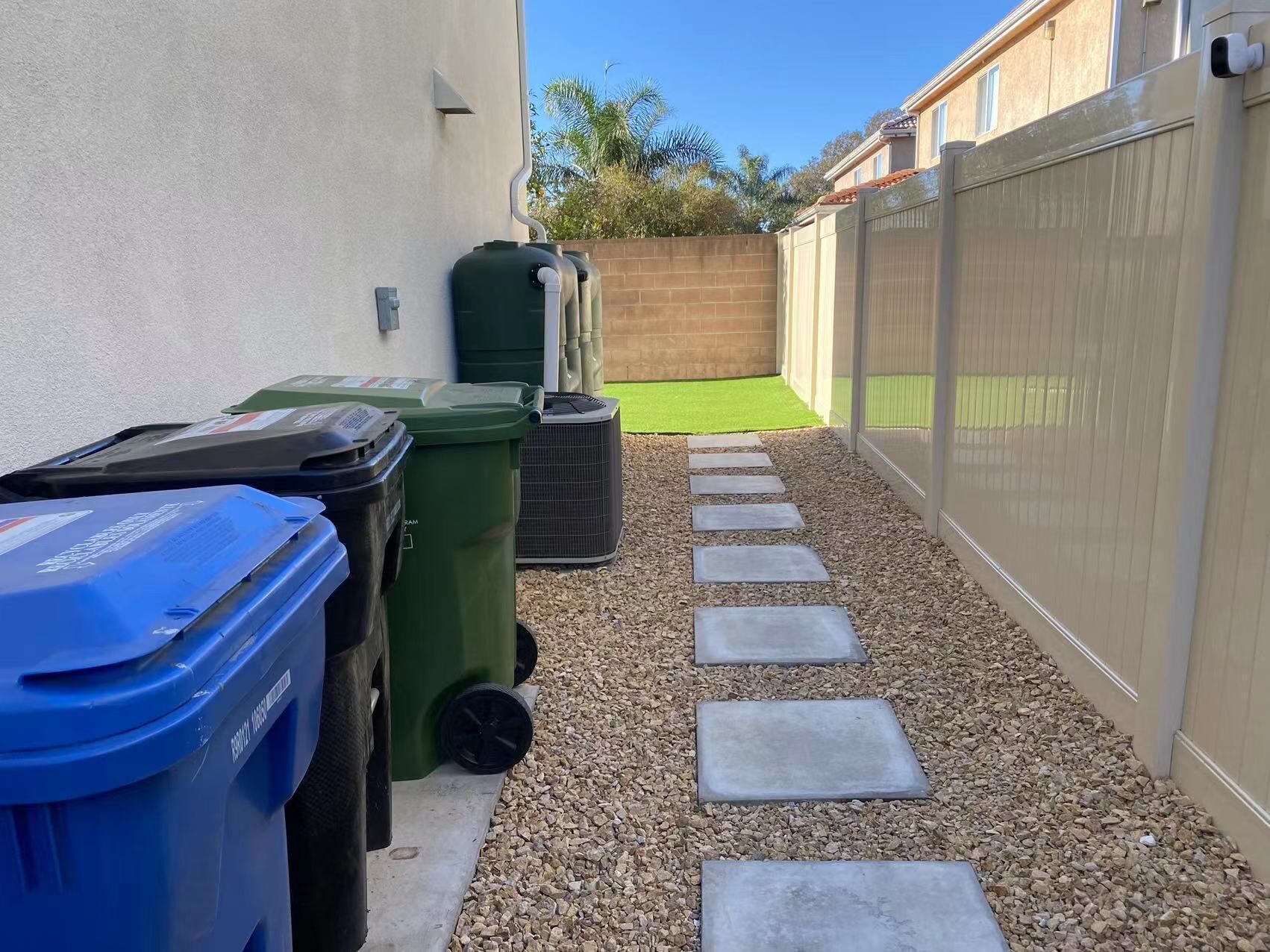 Un étroit sentier extérieur, parsemé de pierres plates pour faciliter le passage, mène à une pelouse verdoyante, bordée de poubelles et d’une clôture.