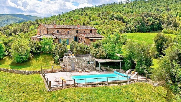 A rustic stone house with a swimming pool, surrounded by lush greenery and hills.