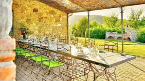 An outdoor dining area with a stone wall backdrop, a covered section with a woven roof, and a view of greenery and hills in the distance.