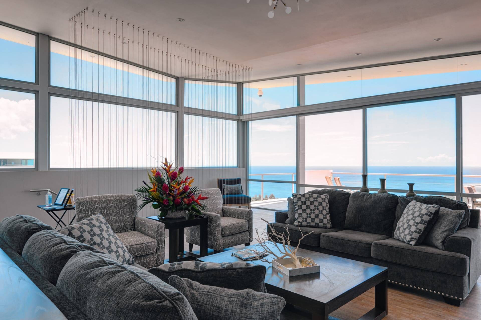 Ein geräumiges Wohnzimmer mit großen Fenstern und Meerblick. Es gibt verschiedene Sitzgelegenheiten, darunter eine Couch und Sessel.