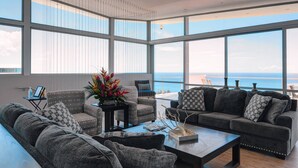En rummelig stue med store vinduer der giver udsigt over havet. Der er flere siddepladser, heriblandt en sofa og lænestole.
