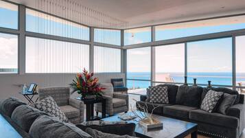 Ein geräumiges Wohnzimmer mit großen Fenstern und Meerblick. Es gibt verschiedene Sitzgelegenheiten, darunter eine Couch und Sessel.