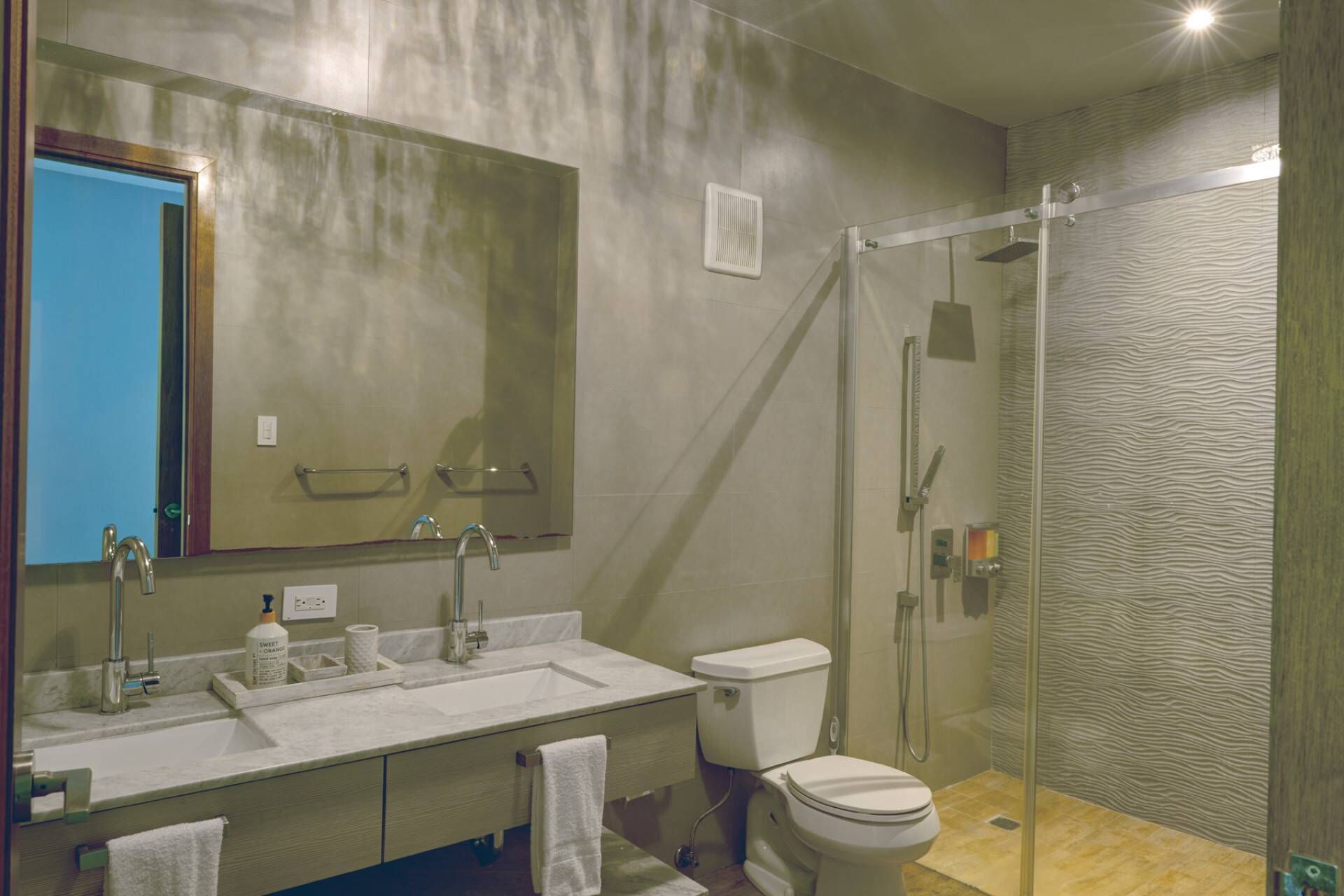A modern bathroom with a large mirror, a sink with a soap dispenser, a toilet, and a glass-enclosed shower area.