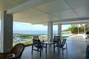 A modern outdoor patio with a view of the ocean, featuring a pool, lounge chairs, and a dining area. - 5BR Villa | Private Pool + Walk-Out Kitchen | Near Seven Seas Beach | Sleeps 14 (Fajardo)