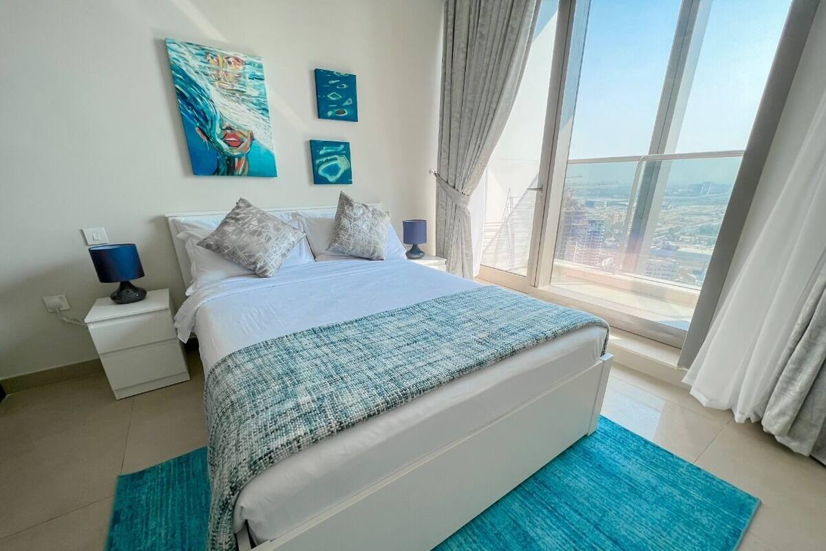 Slaapkamer met een groot bed, nachtkastje, lamp en uitzicht op de stad door een groot raam.