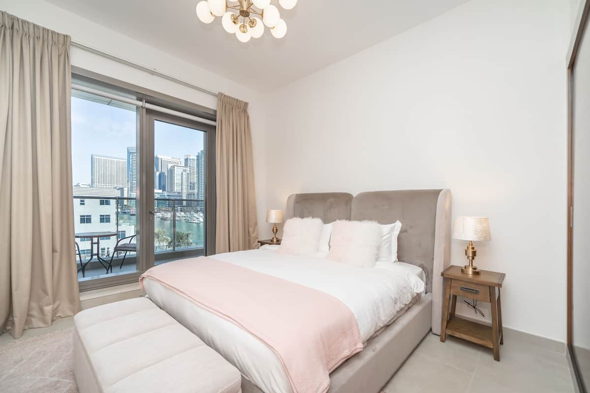Een moderne hotelkamer met een groot bed, een nachtkastje met een lamp en uitzicht op de stad door een groot raam.