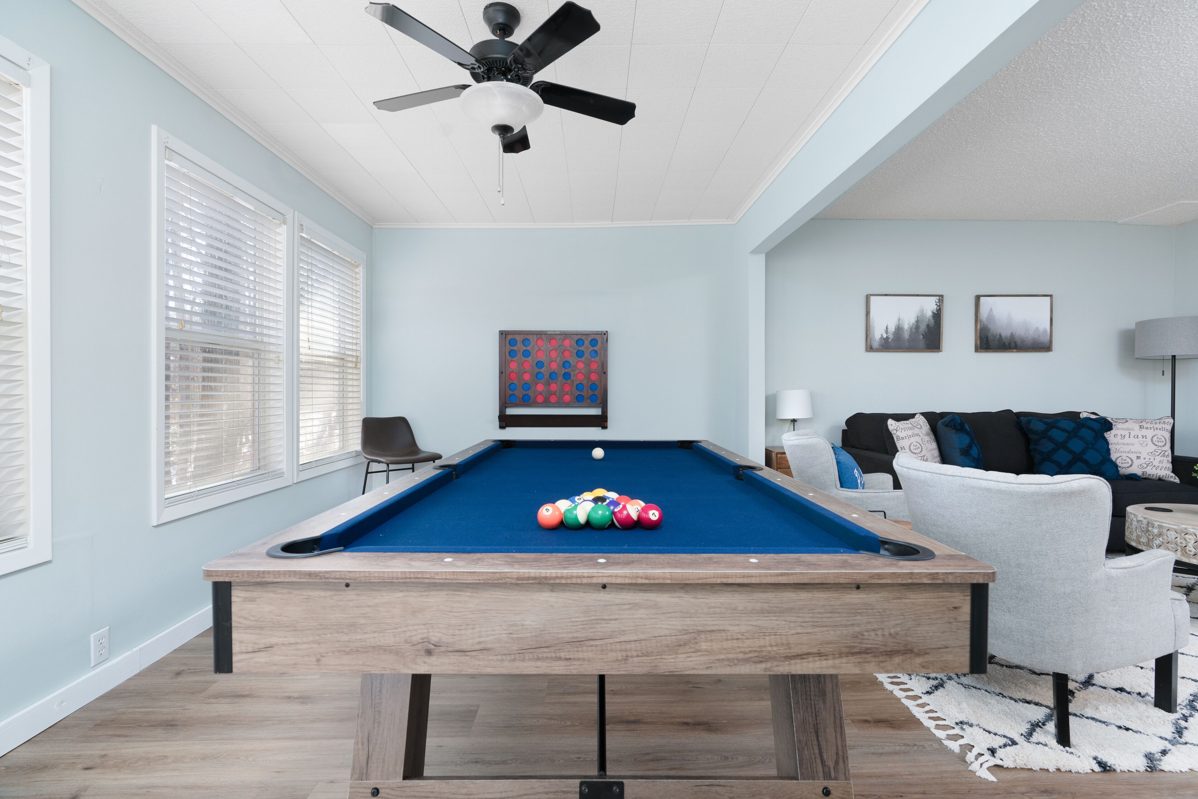 Une table de billard avec des billes, dans une pièce aux murs bleu clair, avec un ventilateur au plafond et un canapé.