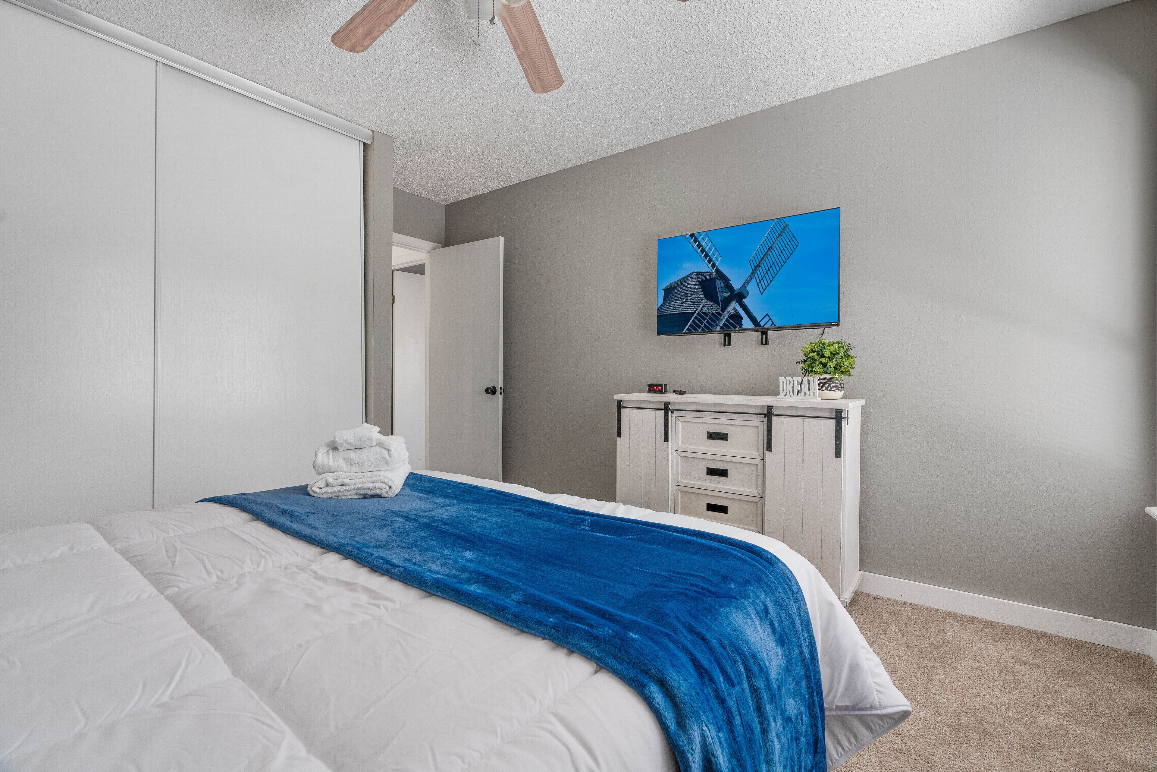 Un dormitorio con cama, manta azul, armario blanco, cómoda y televisor montado en la pared mostrando un molino.