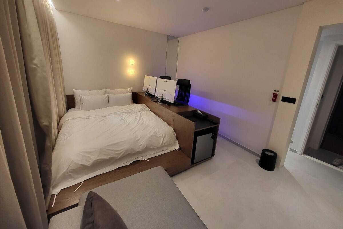 A modern hotel room with a large bed, bedside tables, a desk with a computer, and a small trash bin.