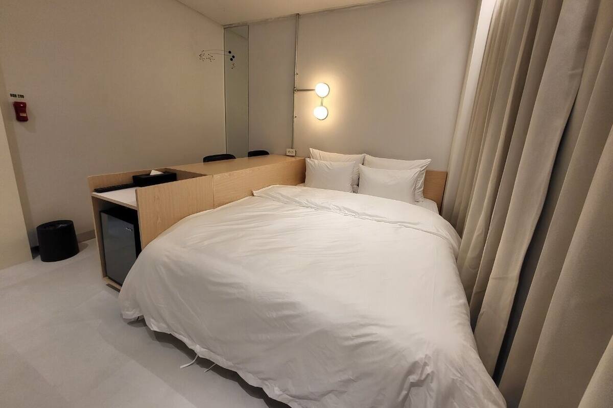 A hotel room with a bed, a desk, a mirror, and a wall-mounted light.