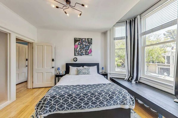 A bedroom with a bed, a window with curtains, a painting on the wall, and a door.
