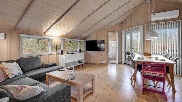 A spacious living area with a sofa, coffee table, and dining table with chairs. A TV is mounted on the wall, and there are large windows providing natural light. - 3 bedroom cozy home in Højslev (Højslev)