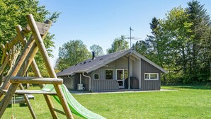 A wooden play structure with a slide, a small house with a sloped roof, and a grassy yard. - 3 bedroom cozy home in Højslev (Højslev)