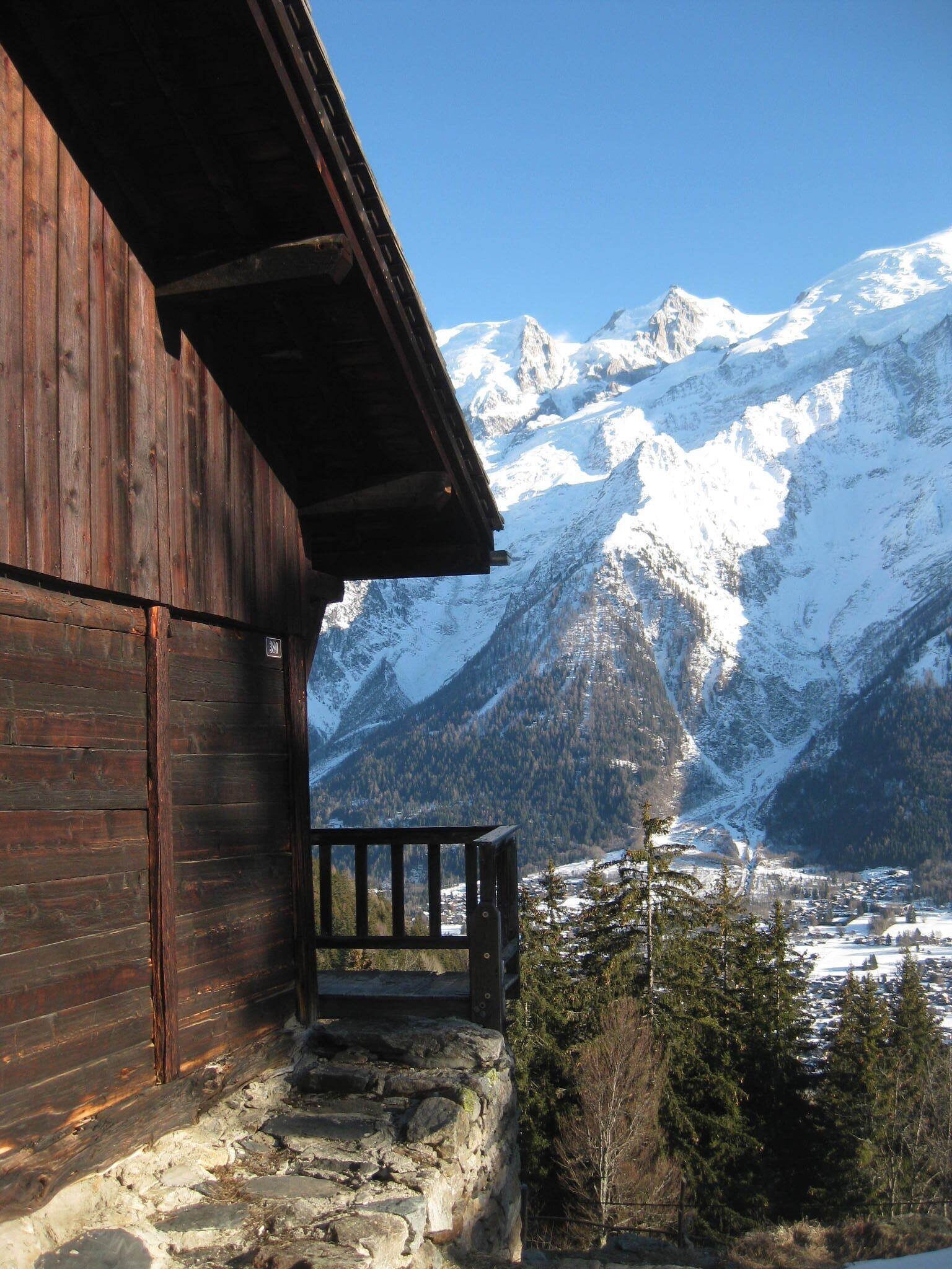 A wooden cabin with a stone foundation, a balcony, and a view of snow-covered mountains.