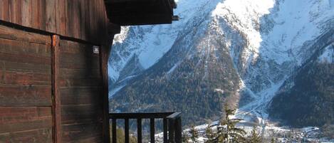 A wooden cabin with a stone foundation, a balcony, and a view of snow-covered mountains.