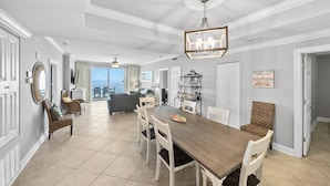 A spacious dining area with a large wooden table and white chairs, a chandelier, and a view to the outside. - Ocean Ritz 1503 • Luxury Beachfront Retreat (Panama City Beach)