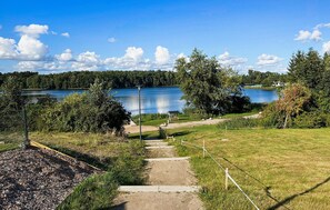 Ein Weg führt zu einem See mit einer Bank und einem Pavillon, umgeben von Bäumen und einem Zaun.