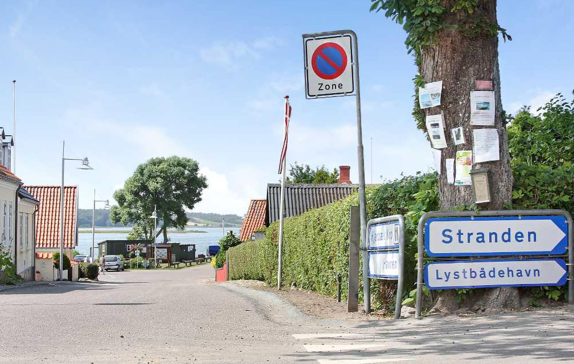 Une route avec un panneau indiquant « accès interdit », un arbre sur lequel sont accrochés des papiers, et des panneaux de signalisation indiquant la direction de « Stranden » et de « Lystbådehavn ».