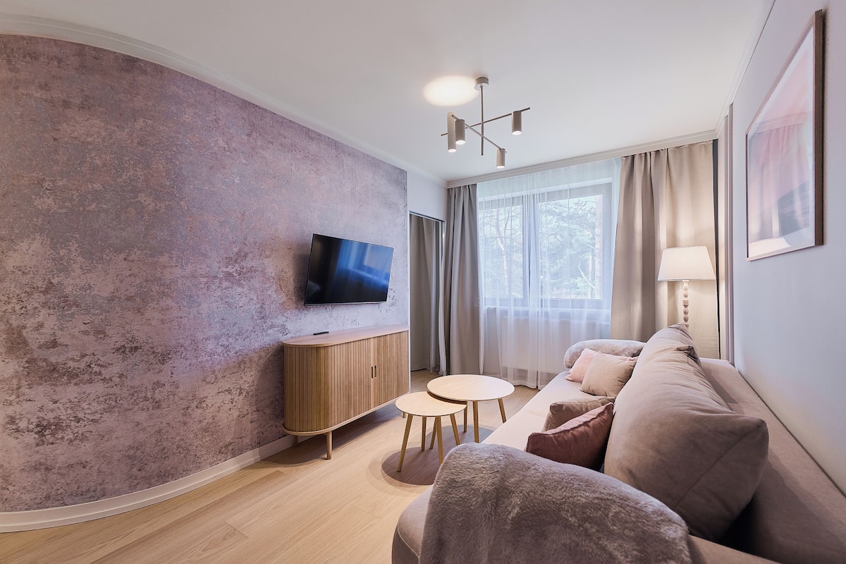 Ein modernes Wohnzimmer mit Flachbildfernseher, Holzschrank, rundem Couchtisch, beiger Couch und großem Fenster mit Vorhängen.