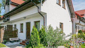 A two-story house with a white exterior, red-tiled roof, and multiple windows. There is a small garden area with green shrubs and a bicycle parked outside. - 2 bedroom amazing apartment in Bobolin (Bobolin)