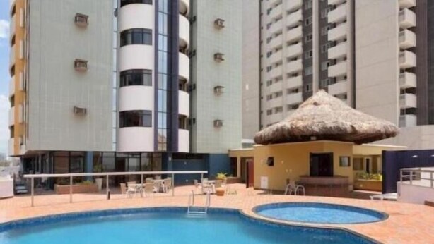 A swimming pool with a thatched roof structure, surrounded by a poolside area with chairs and a small building. - Silva Hospedagem - Flat Number One (São Luís)