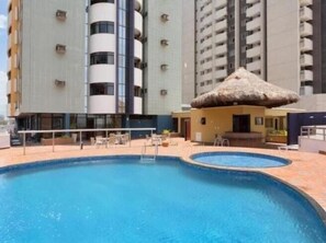 A swimming pool with a thatched roof structure, surrounded by a poolside area with chairs and a small building. - Silva Hospedagem - Flat Number One (São Luís)