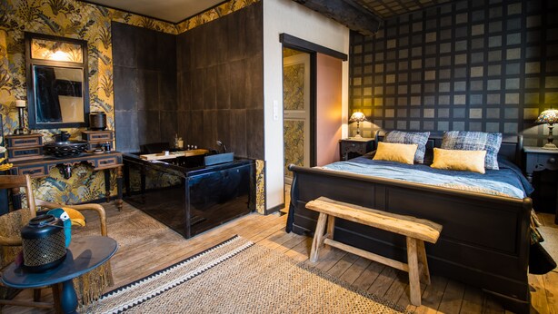 A bedroom with a large bed, a wooden bench, a desk with a chair, a vanity with a mirror, and a bathroom with a sink and mirror. - Unique cosy Petite Maison with luxury jacuzzi (Hesdin)