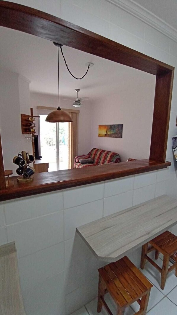 A kitchen with a wooden countertop, a hanging pendant light, a ceiling fan, and a window with a view of a room with a sofa.