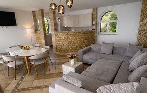 Una sala de estar moderna con un sofá gris, una mesa de comedor con sillas, una zona de cocina con paredes de piedra y un televisor montado en la pared.