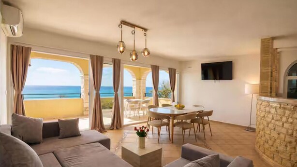 A living room with a sofa, a dining table with chairs, a TV mounted on the wall, and a view of the sea through large windows. - Lovely home in Jesenice with sauna (Jesenice)