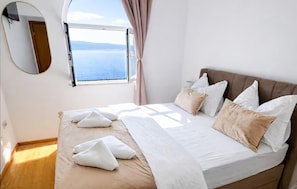 A bedroom with a bed, pillows, a window with a view, a mirror, and a wooden floor.
