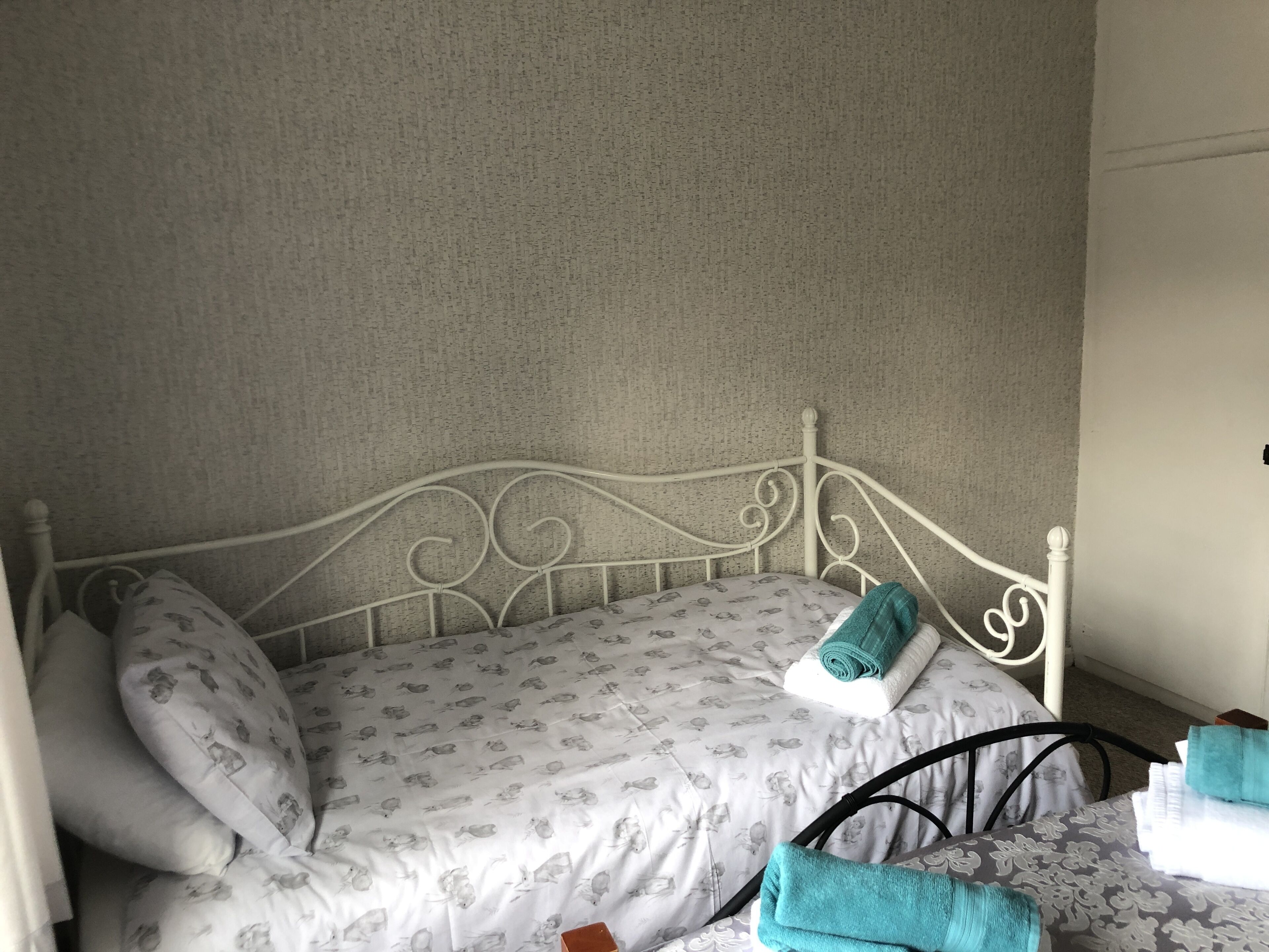 Una cama individual con estructura metálica blanca, ropa de cama estampada y una toalla enrollada en la cabecera.