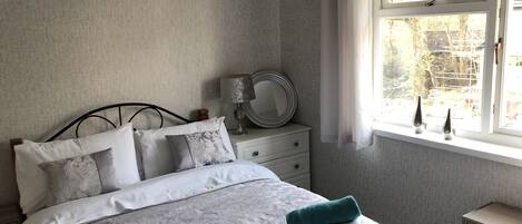 A neatly made bed with a white and grey patterned duvet, two teal towels, a bedside table with a lamp and a mirror, and a window with a view of the outdoors.