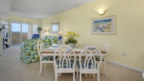 A dining area with a table set for four, white chairs, and a painting on the wall. - Playa Flamingo 104-Bayside 83rd St, Free WiFi, Pool, Elev, W/D, AC (Ocean City)