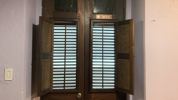 Een houten dubbele deur met een 'No Smoking' bord erboven, aan weerszijden omlijst door twee ramen met zonwering.