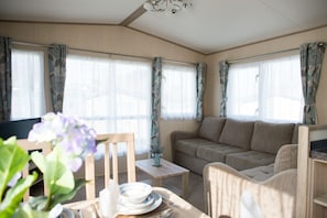 A cozy living room with a sofa, a small table, and a chair. There are curtains on the windows, and a plant is visible on the table.