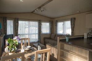 A cozy living room with a sofa, dining table, and chairs. There are large windows with curtains, a ceiling fan, and a small kitchen area with a counter and sink.