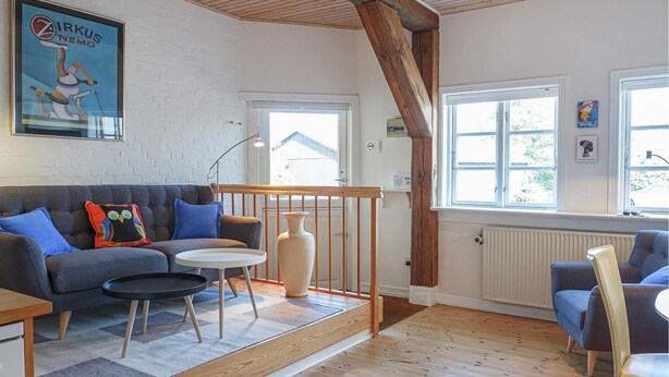 Een gezellige woonkamer met een bank, een klein rond tafeltje en een stoel. Er is een houten trap met een leuning, een raam met uitzicht en een radiator.