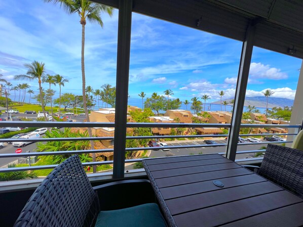 A balcony with wicker furniture, a view of a coastal area with palm trees and buildings, and a clear sky.