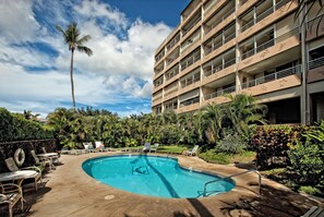 A swimming pool with lounge chairs and a palm tree, surrounded by greenery and a multi-story building.