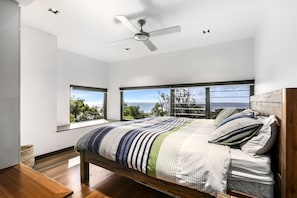 Quarto com cama, ventilador de teto e grandes janelas com vista para o mar.
