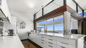 A modern kitchen with a large island, white cabinetry, and a view of the sky through large windows. - Banyandah - Luxury Architect Designed Straddie Beachhouse (Point Lookout)